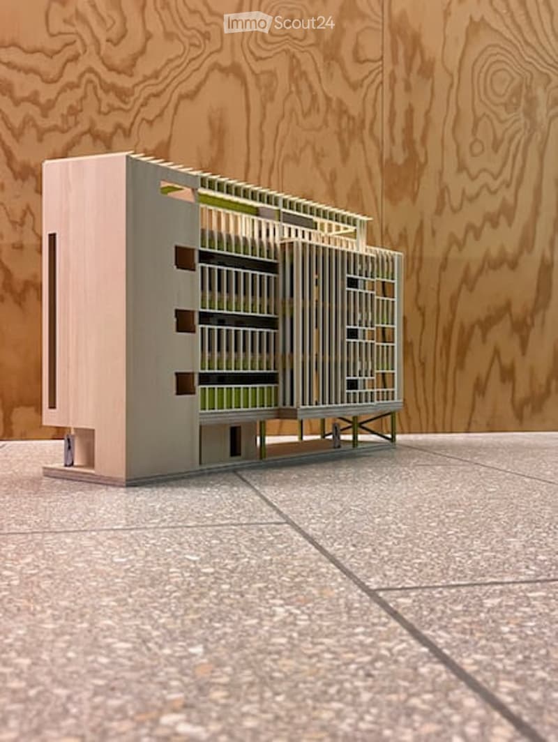 A modern, minimalist building model with a clean geometric design, featuring a combination of light pink and grey materials, set against a wooden backdrop.