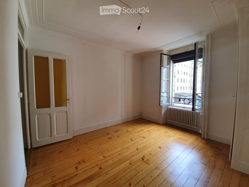 Empty room with wooden flooring, white walls, and a large window with a radiator below it. The room has a door on the left side and a visible light fixture on the ceiling.