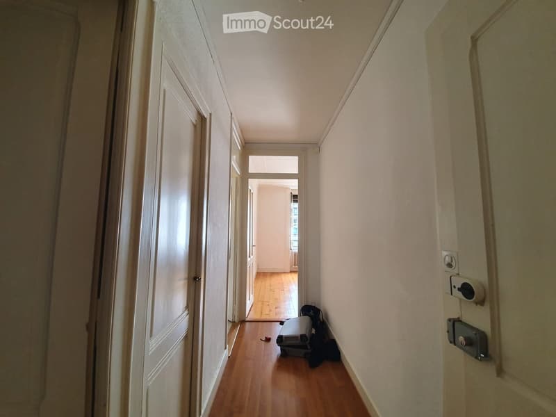 A narrow hallway with wooden flooring, white walls, and several doors on the left side. A small area at the end shows a glimpse of another room with a radiator and a small piece of furniture.