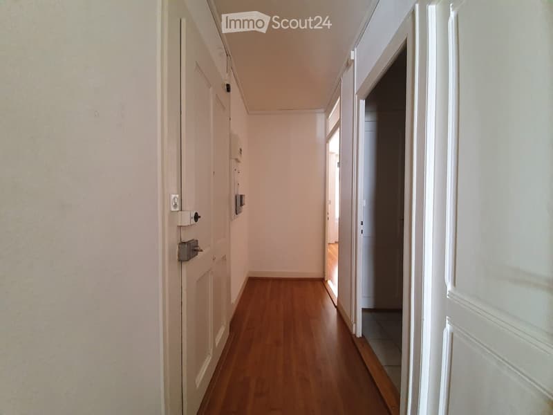 A narrow hallway with wooden flooring and white walls, featuring several doors and a visible light switch on the left side.
