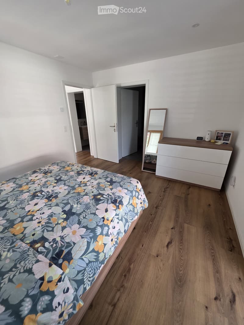 Bright bedroom with wooden flooring, a bed with a floral duvet, a dresser with a mirror, and a picture frame on top.