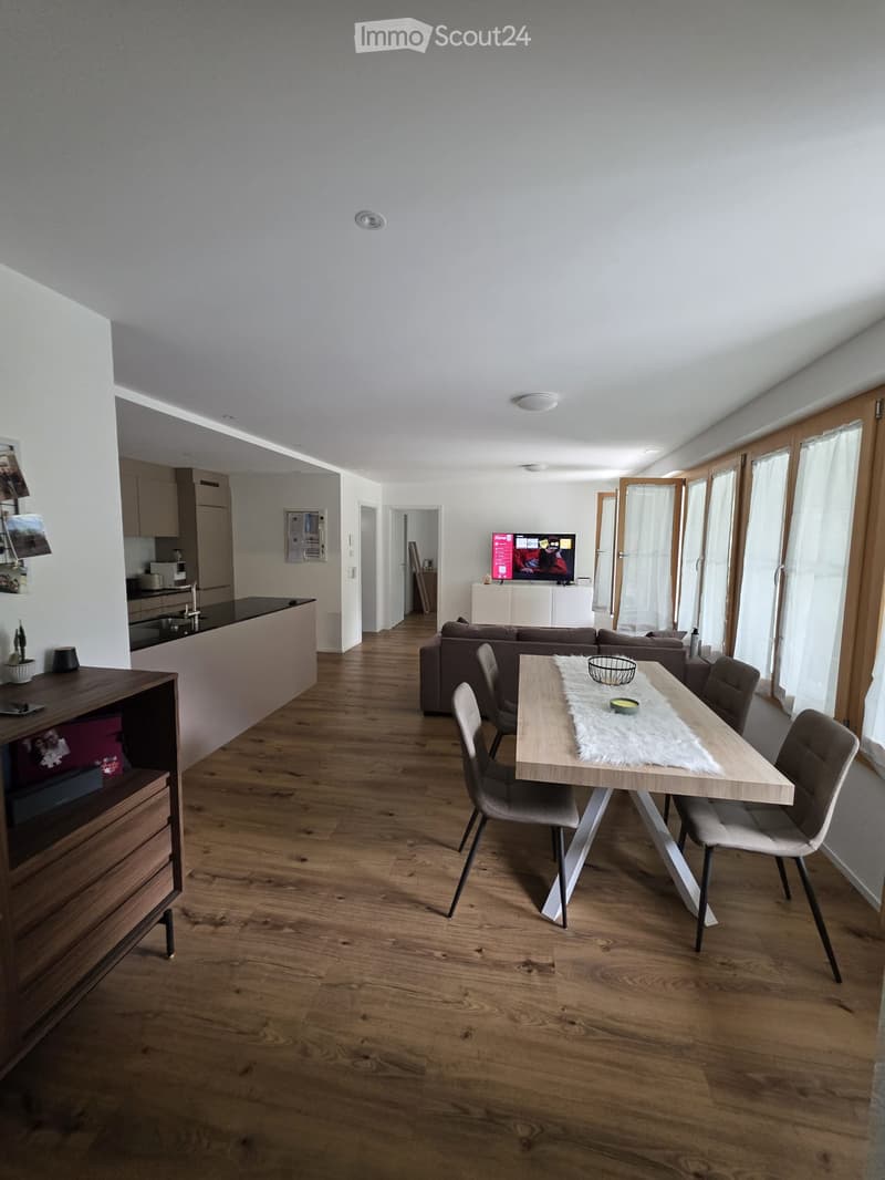 Open-plan living and dining area with wooden flooring, a sofa, a dining table with chairs, a kitchen counter, and a television. Large windows and modern decor.