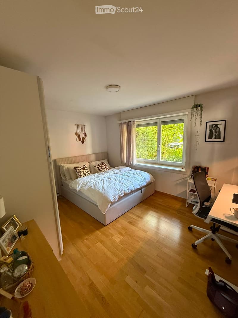Cozy bedroom with a large bed, wooden floor, desk, chair, and decorative items including picture frames and a wicker basket.