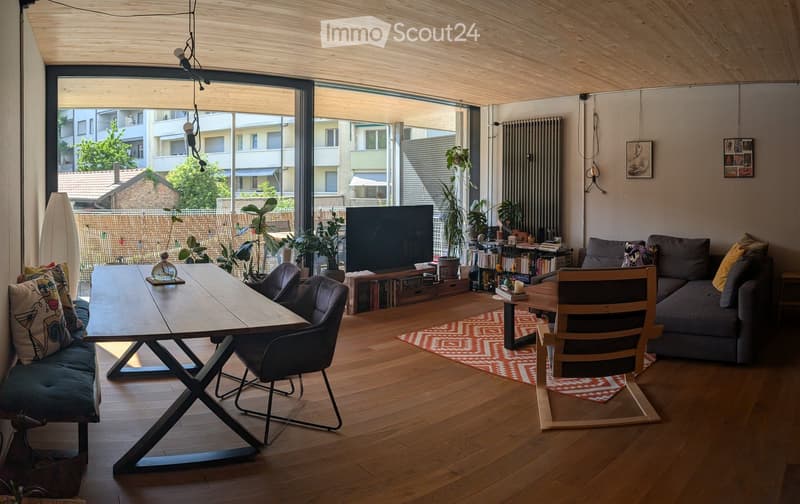 Modern living room with wooden flooring, large windows, a grey sofa, a wooden dining table, chairs, a television, and decorative plants.