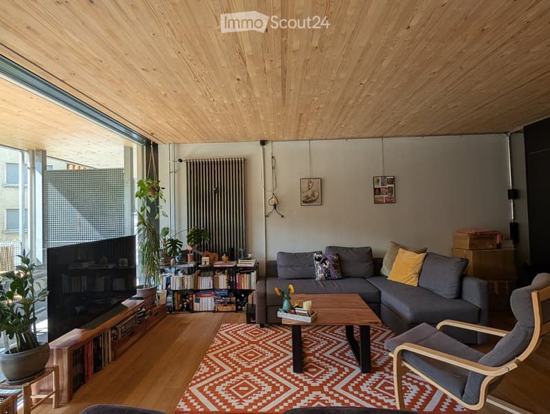Modern living room with wooden ceiling, large window, grey sectional sofa, wooden coffee table, armchair, bookshelf, and plants.