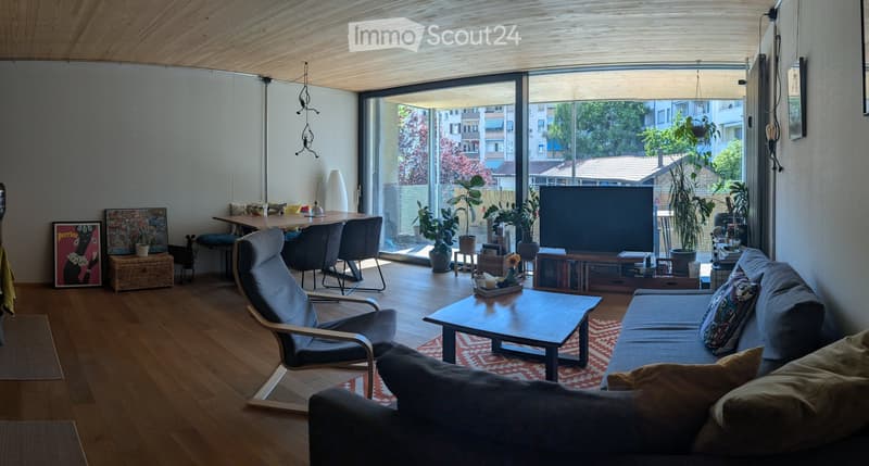 Modern living room with wooden floors, large windows, a grey sofa, coffee table, chairs, dining table, TV, and several potted plants.