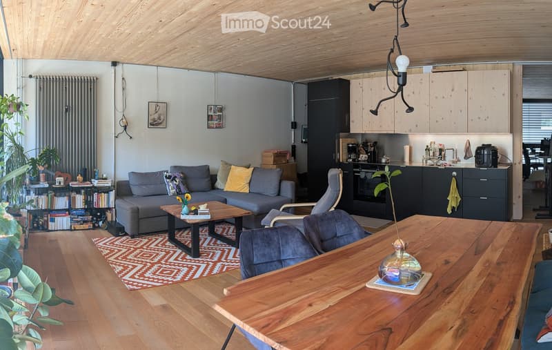 Open-plan living and dining area with wooden floors, grey sofa, coffee table, dining table, chairs, bookshelf, kitchen area with cabinets, oven, and fridge.