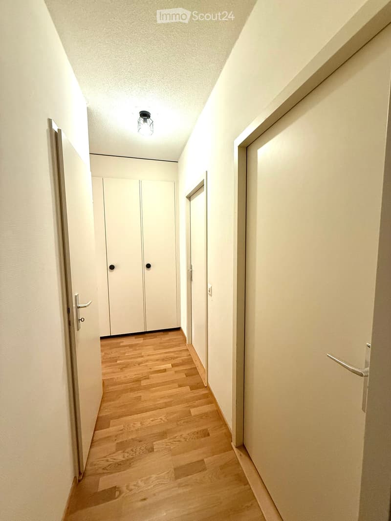 A narrow hallway with light wooden flooring, white walls, and several closed doors. A single ceiling light fixture is visible.