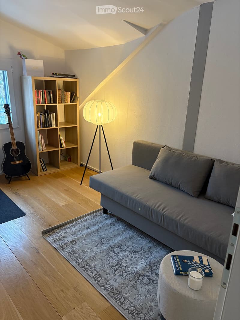 Cozy living room with a grey sofa, wooden bookshelf, guitar, floor lamp, and a small round stool with books and a candle.