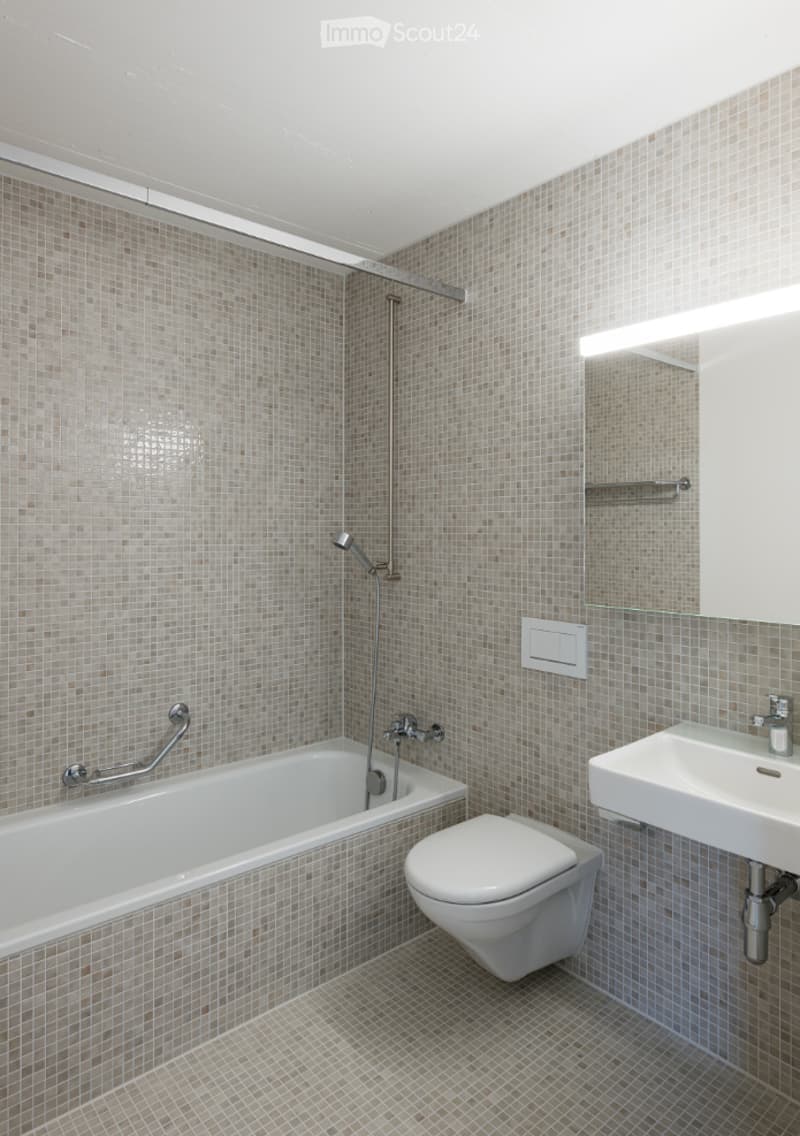 Modern bathroom with beige tiled walls and floor, featuring a bathtub with a handheld shower, a toilet, a sink with a mirror, and a built-in shelf above the sink.