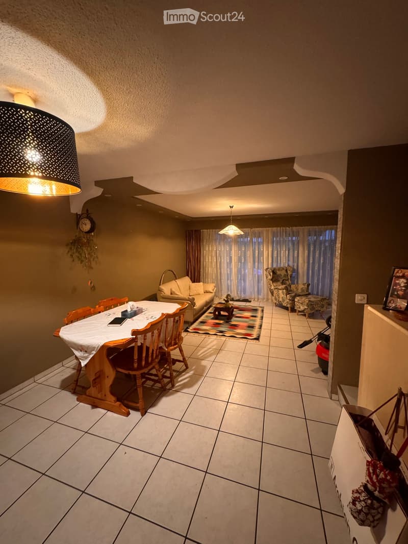 Dining and living area with tiled floor, wooden dining table with chairs, sofa, armchair, coffee table, and decorative rug.