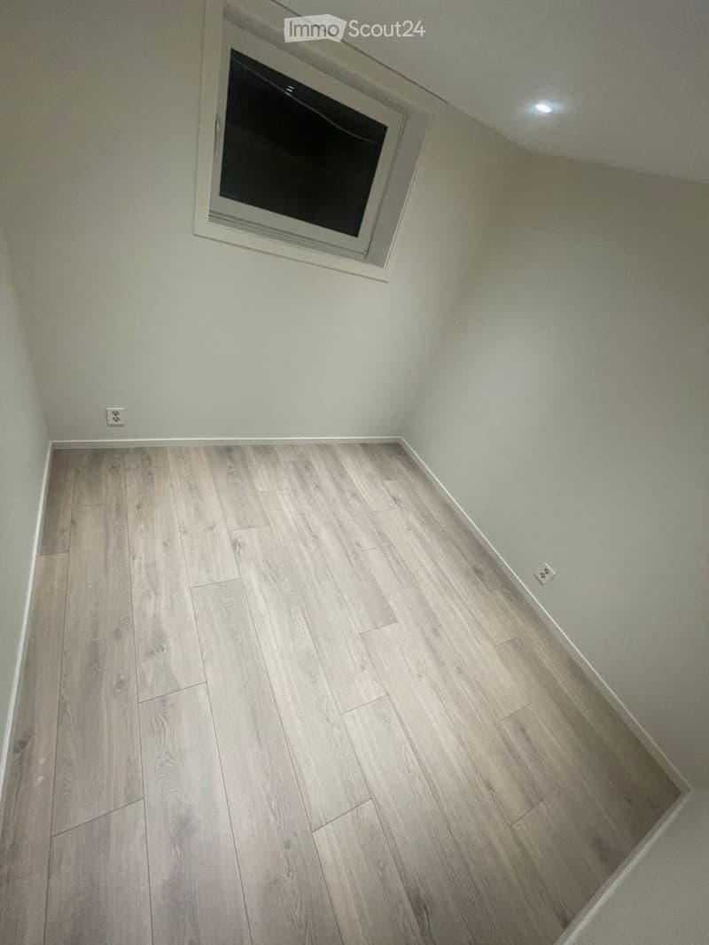 Empty room with light grey walls and light wood flooring, featuring a small window with a white frame and a recessed ceiling light.
