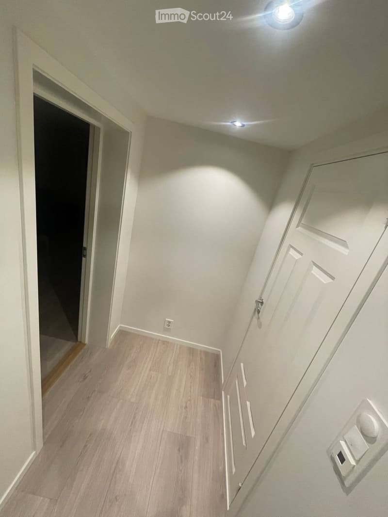 Empty hallway with light wood flooring, white walls, and recessed ceiling lights. Features a closed white door and an open doorway leading to another space.