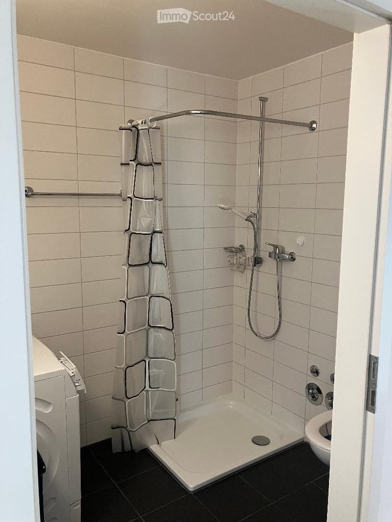 Modern bathroom with a walk-in shower, toilet, and a shower curtain. The shower features a handheld sprayer and a built-in shelf. The walls are tiled in light beige, and the floor is black.