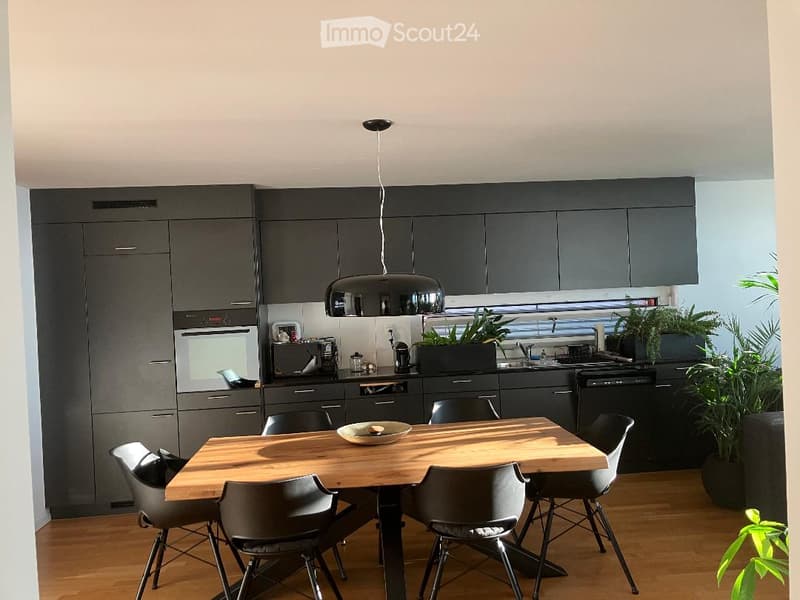 Modern kitchen with grey cabinets, wooden dining table, six chairs, pendant light, and potted plants.