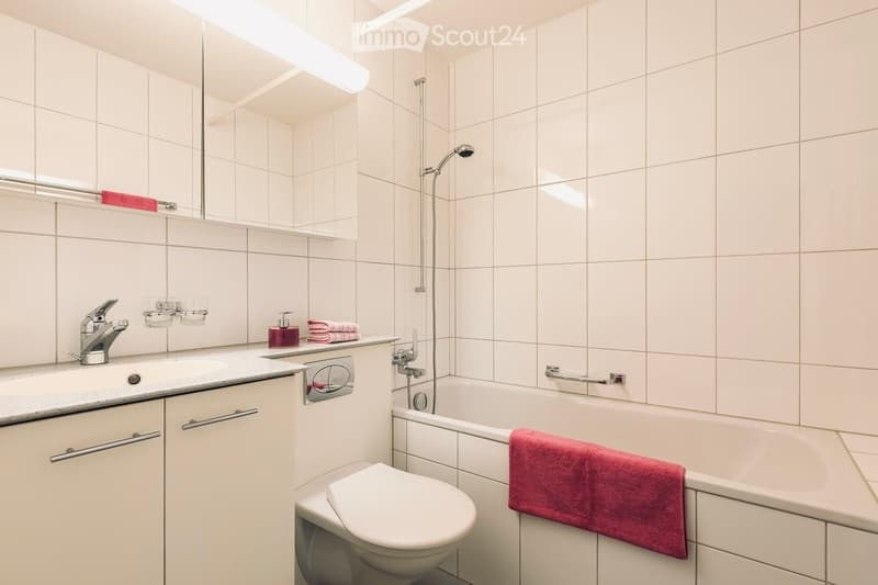 Modern bathroom with white tiles, a bathtub, toilet, sink, and mirror. Includes a red towel on the bathtub and a soap dispenser on the sink counter.