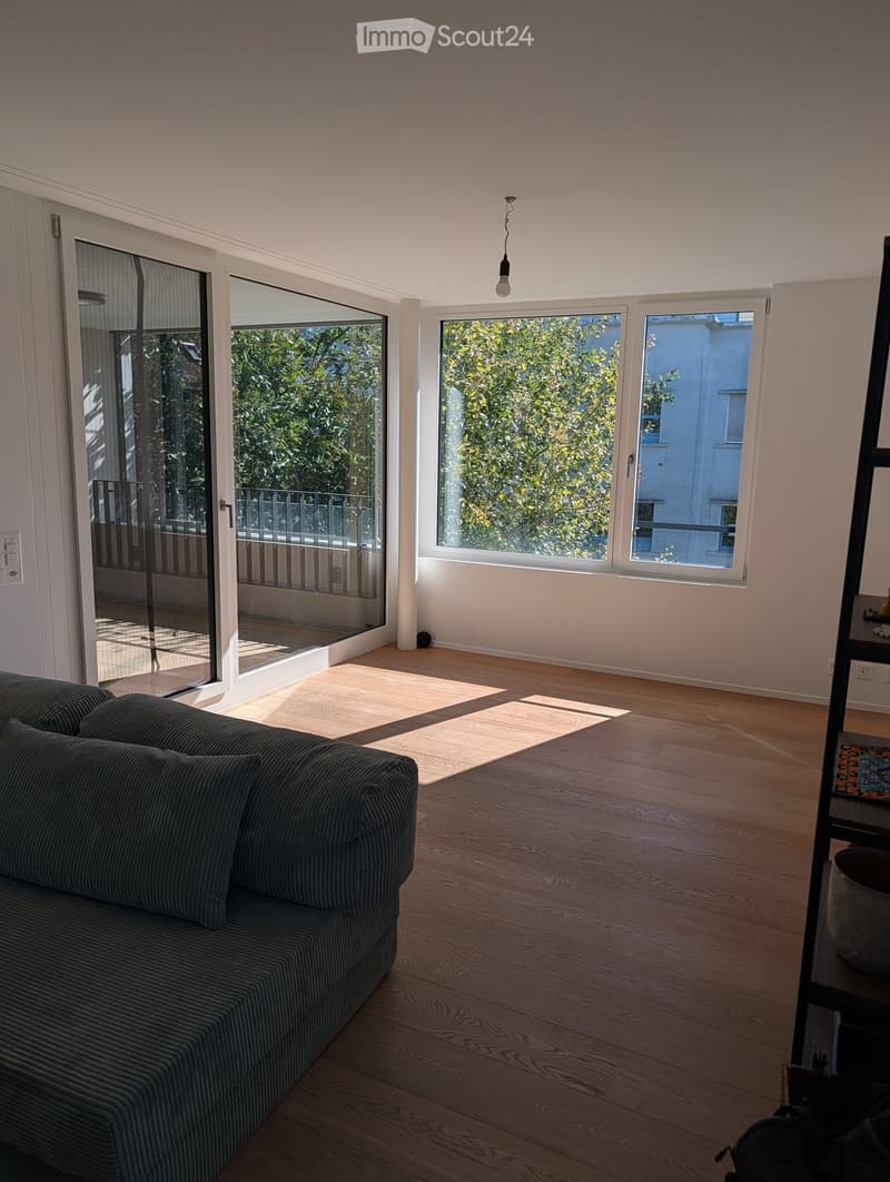 Bright living room with wooden flooring, large windows, and a sliding glass door leading to a balcony. Features a dark grey sofa and a pendant light fixture.