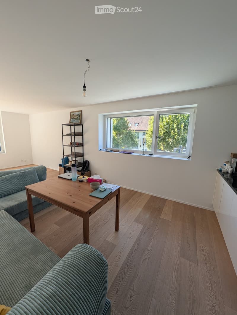 Bright living room with wooden flooring, large window, grey sofa, wooden coffee table, shelf with decorative items, and laptop on table.