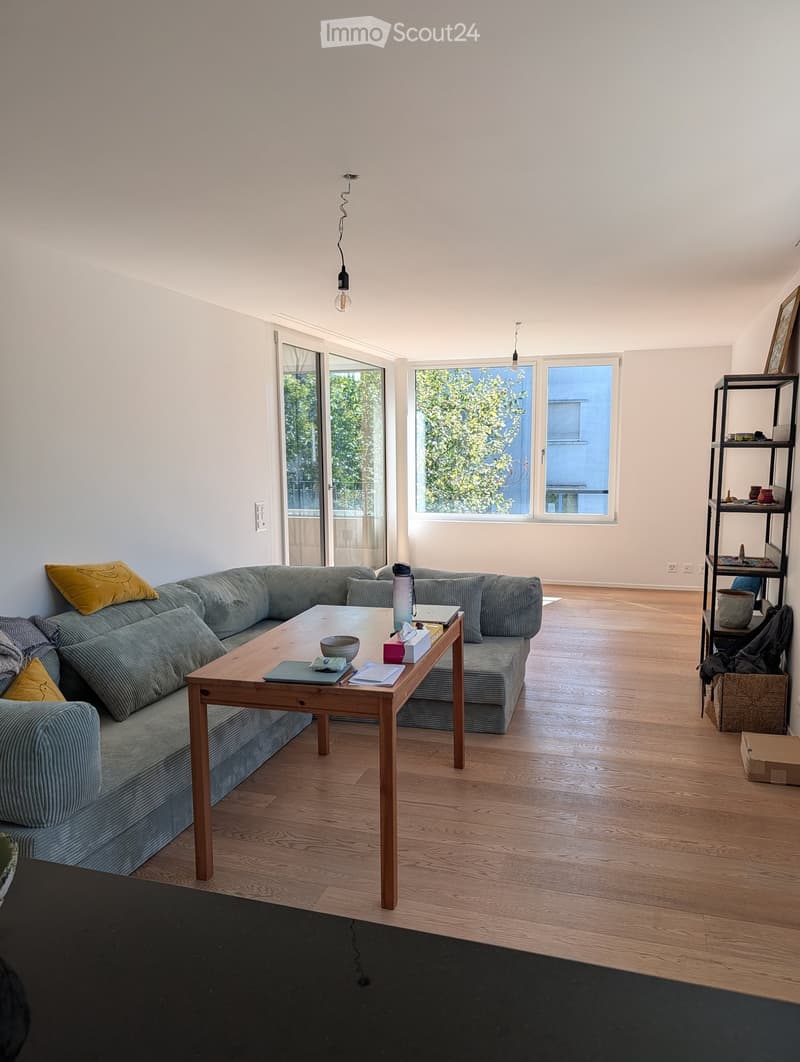 Modern living room with a grey sectional sofa, wooden coffee table, and large windows letting in natural light. The room features a wooden floor and a black shelf with decorative items.
