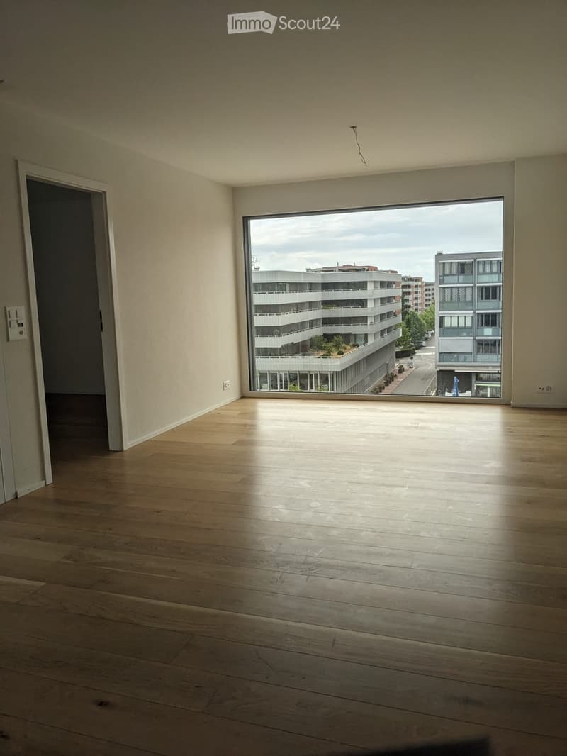 Empty room with wooden flooring, large window showing cityscape, and a door leading to another space.