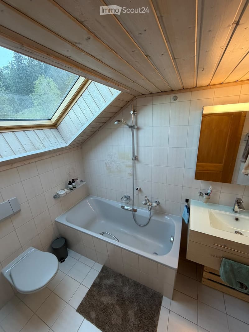 Modern bathroom with a bathtub, toilet, sink, mirror, and wooden cabinet. Features a skylight window and tiled walls.