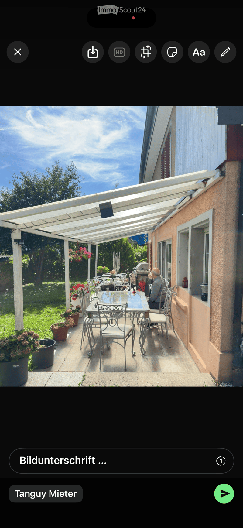 A covered outdoor patio with a glass table, white metal chairs, and potted plants, adjacent to a house with a view of a garden.