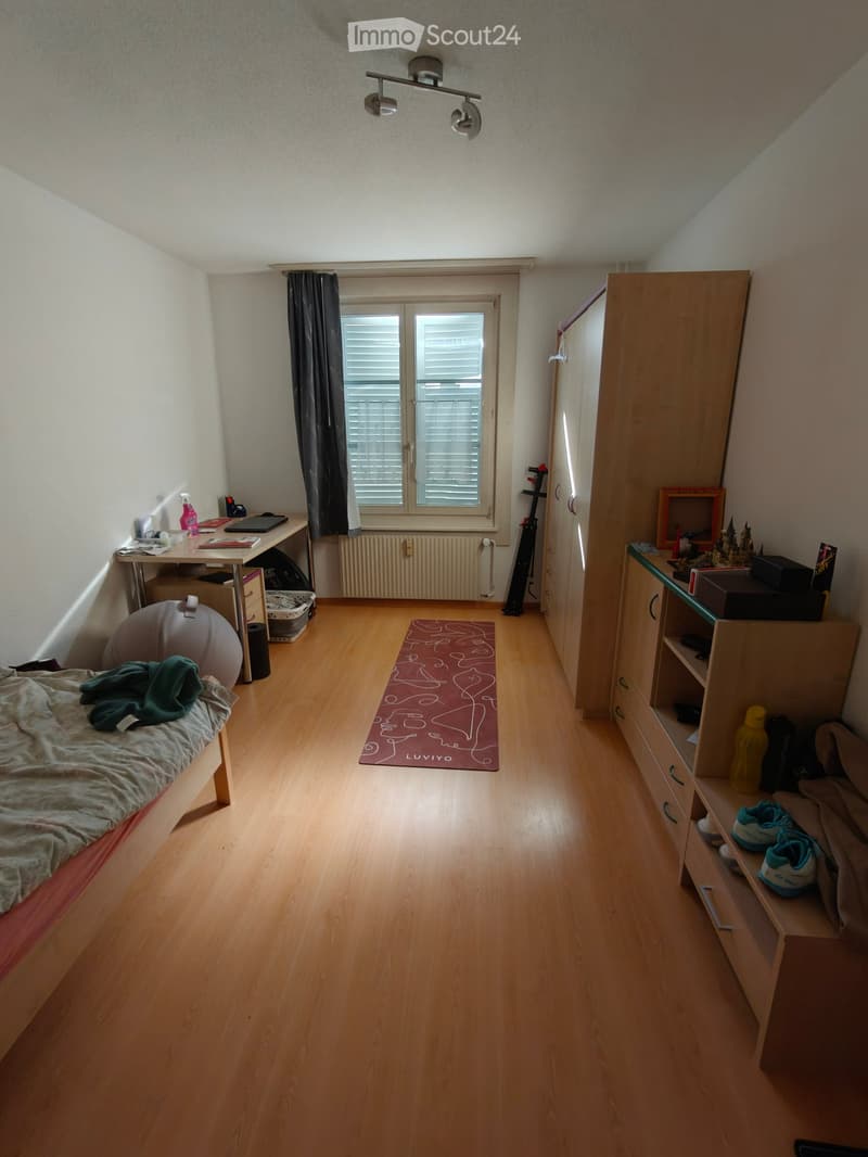 The image shows a furnished bedroom with a bed, desk, wardrobe, shelf, and various personal items. The room features wooden flooring, a window with blinds, and a radiator under the window.