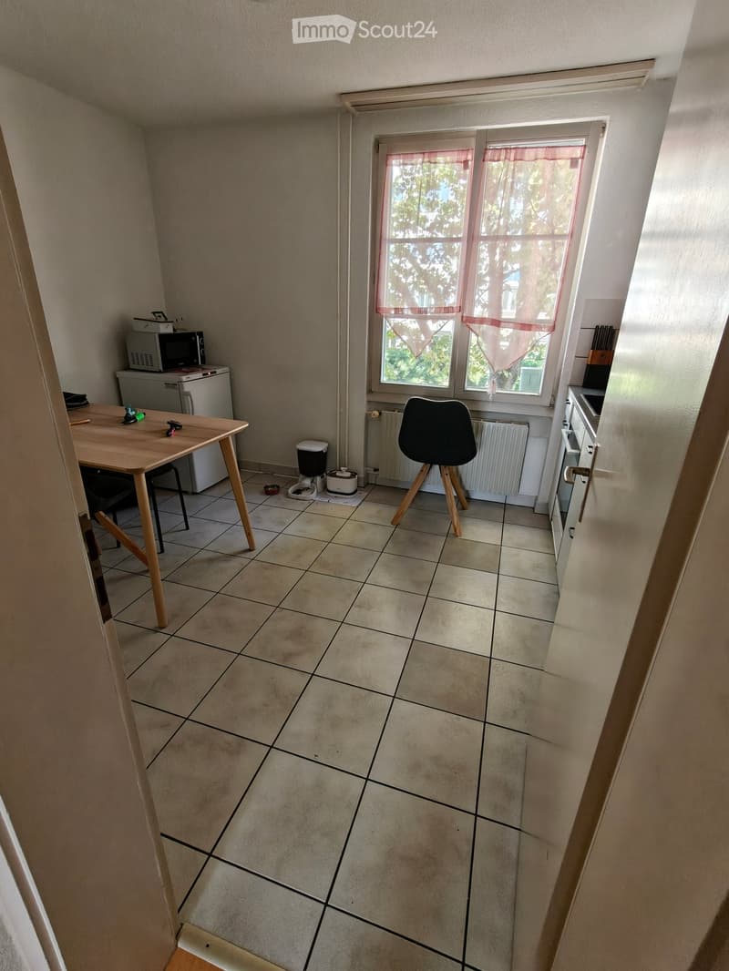 A small kitchen area with tiled flooring, a wooden table, a black chair, a microwave, a refrigerator, and a radiator near the window.