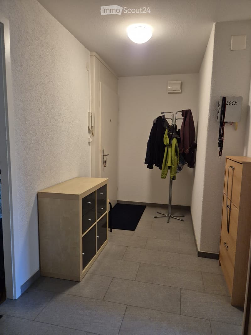 Clean hallway with tiled floor, white walls, coat rack with jackets, small wooden cabinet, and a black doormat near the entrance.