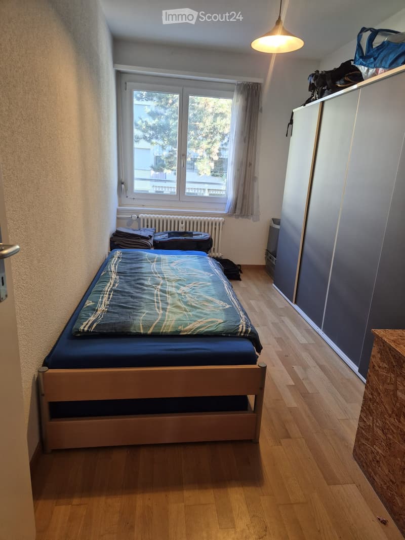 Cozy bedroom with wooden floor, single bed with blue and green bedding, radiator, wardrobe, and window with curtain.