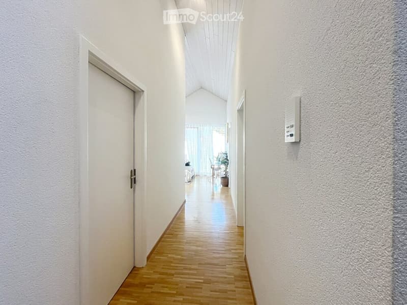 A clean, well-lit hallway with wooden flooring, white walls, and a door on the left leading to another room.