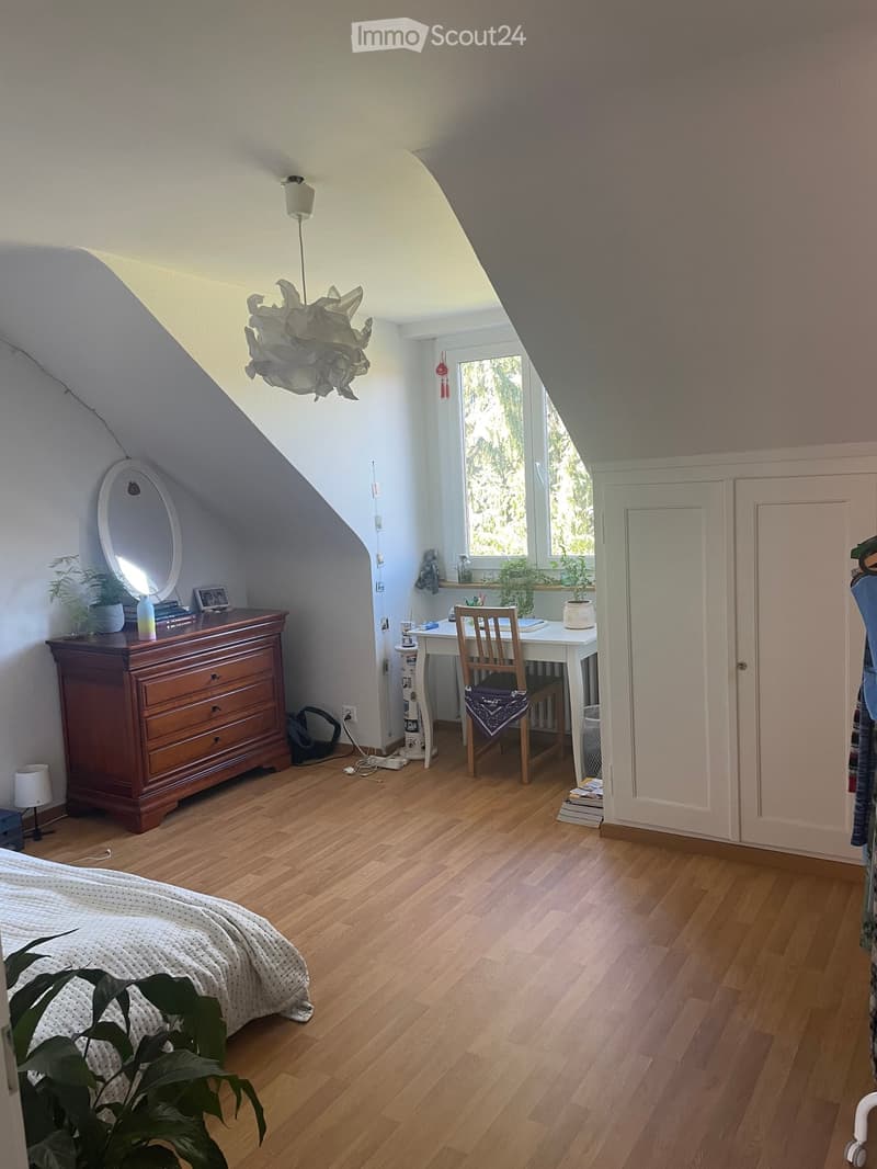Cozy attic bedroom with wooden flooring, slanted ceilings, and large windows. Features a bed, desk with chair, dresser with mirror, and decorative plants.