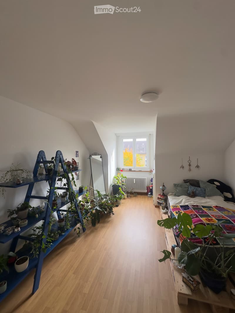 Cozy attic bedroom with wooden flooring, slanted ceilings, and large window. Features a bed with colorful quilt, a shelf with potted plants, a mirror, and a small table with additional plants.