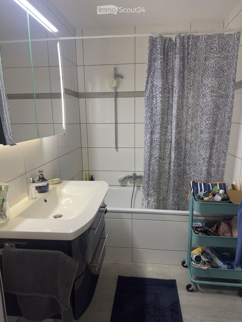 Modern bathroom with white tiles, a sink with a faucet, a bathtub with a shower curtain, a mirror, a towel, and a cart with toiletries.