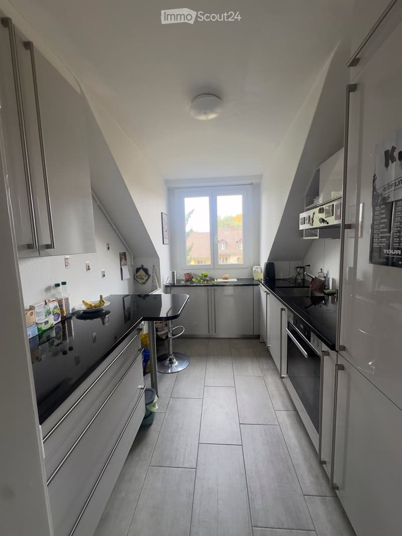 Modern kitchen with stainless steel appliances, grey cabinets, and a black countertop. Features include a refrigerator, oven, sink, and a small bar area with a stool.
