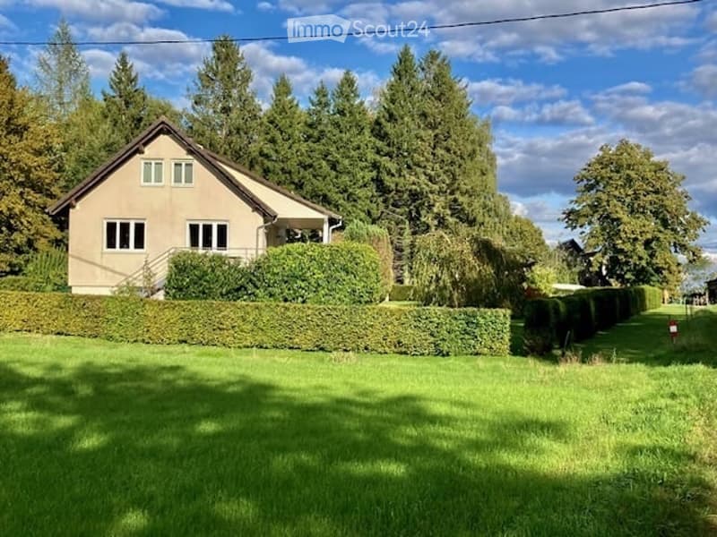 Single-story house with a pitched roof, surrounded by a well-maintained lawn and tall trees. The house has multiple windows and a small porch area.