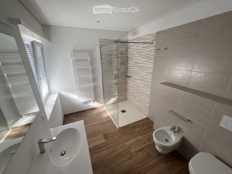 Modern bathroom with wooden flooring, white sink, toilet, bidet, and a glass-enclosed shower area. Features a radiator and a window with blinds.