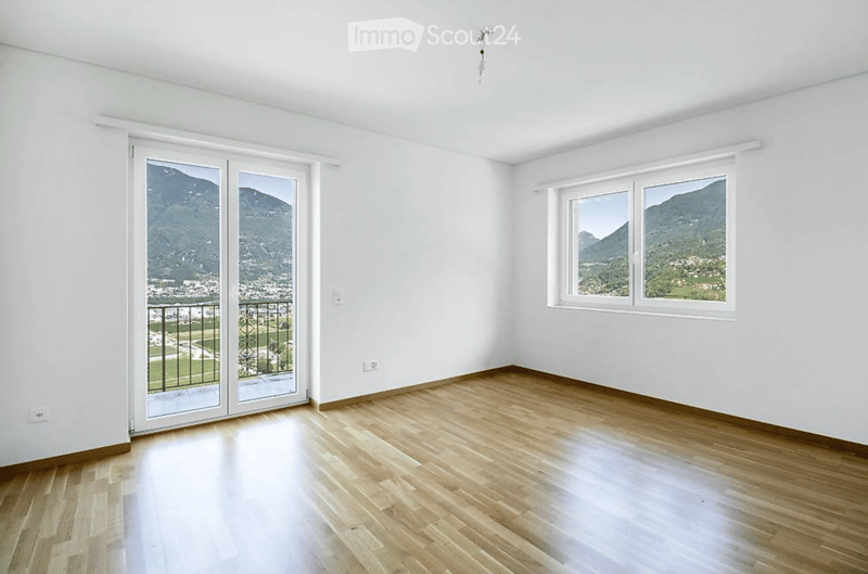 Empty room with wooden flooring, white walls, large windows, and a sliding glass door leading to a balcony with a scenic mountain view.