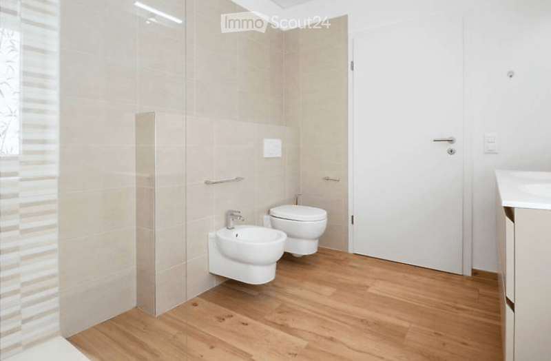Modern bathroom with wooden flooring, white tiles, a toilet, a bidet, and a sink. Features a glass shower enclosure and a white door.