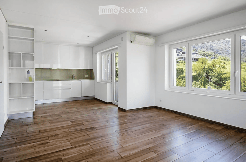 Empty room with white walls, wooden flooring, white cabinets, and large windows offering a scenic view.