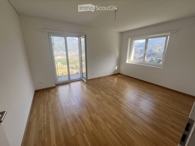 Empty room with wooden flooring, large windows offering a scenic mountain view, and a sliding glass door leading to a balcony.