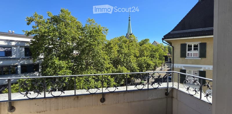 Balcony with metal railing overlooking a street lined with trees and buildings, including a church spire in the distance.