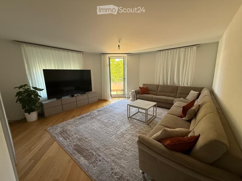 Modern living room with a large sectional sofa, coffee table, flat-screen TV, and a potted plant. The room features wooden flooring, large windows with white curtains, and access to a balcony.
