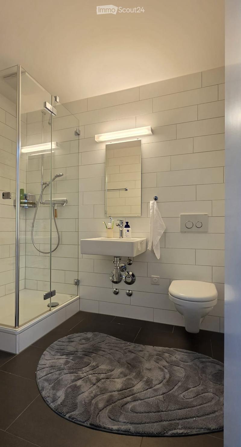 Modern bathroom with white tiles, a glass-enclosed shower, a white sink with a mirror, a toilet, and a soft grey round rug on the dark floor.