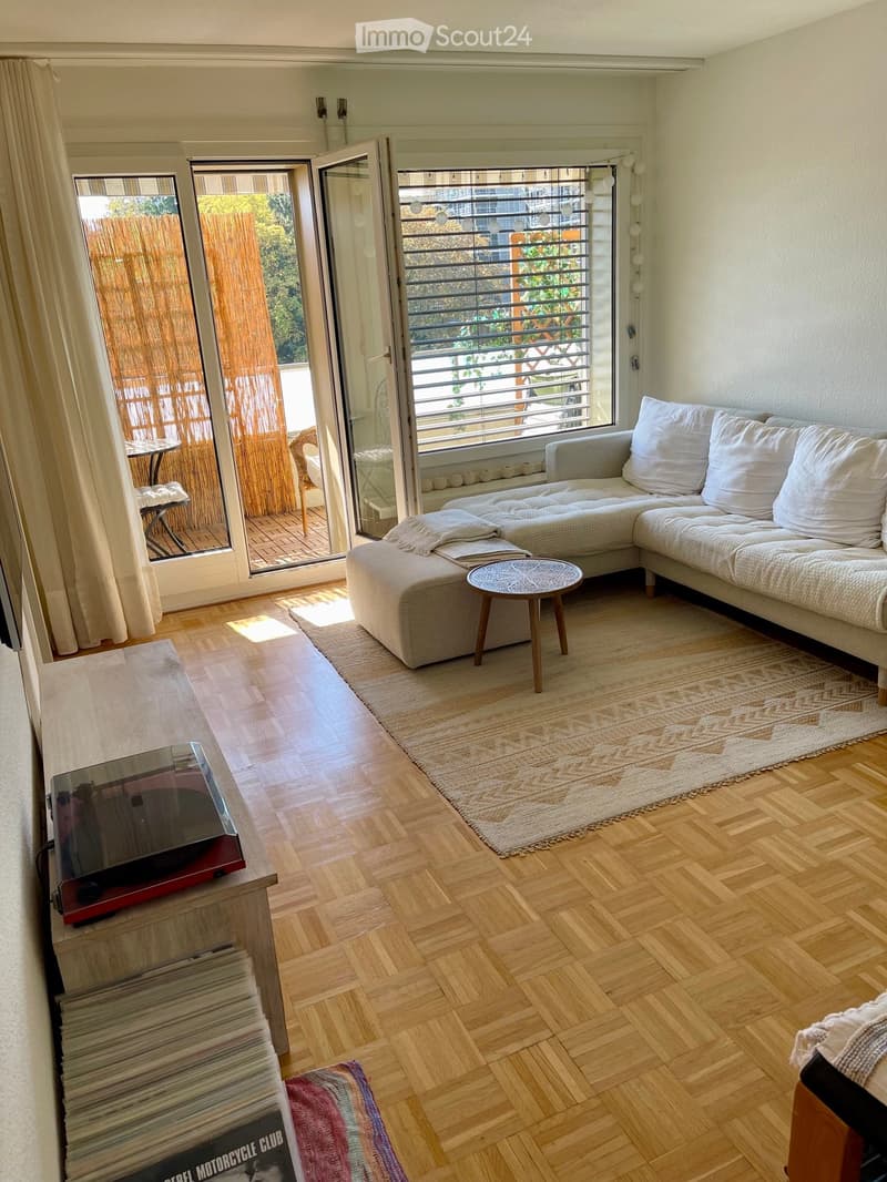 Living room with a grey sectional sofa, coffee table, TV, and a small table and chair set near the balcony. Wooden flooring and large windows with a view of the balcony.