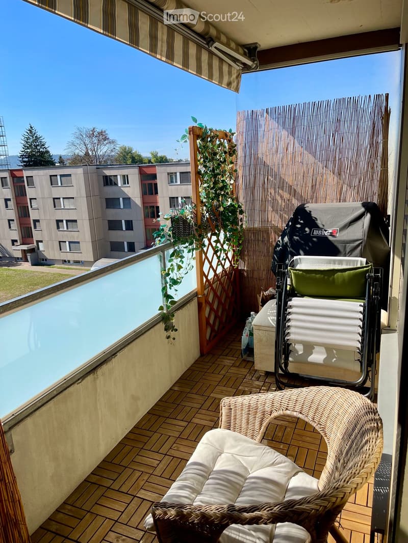 Balcony with wooden decking, wicker chair, sun lounger, barbecue, and climbing plants on a wooden trellis. The view shows a multi-story building and greenery.