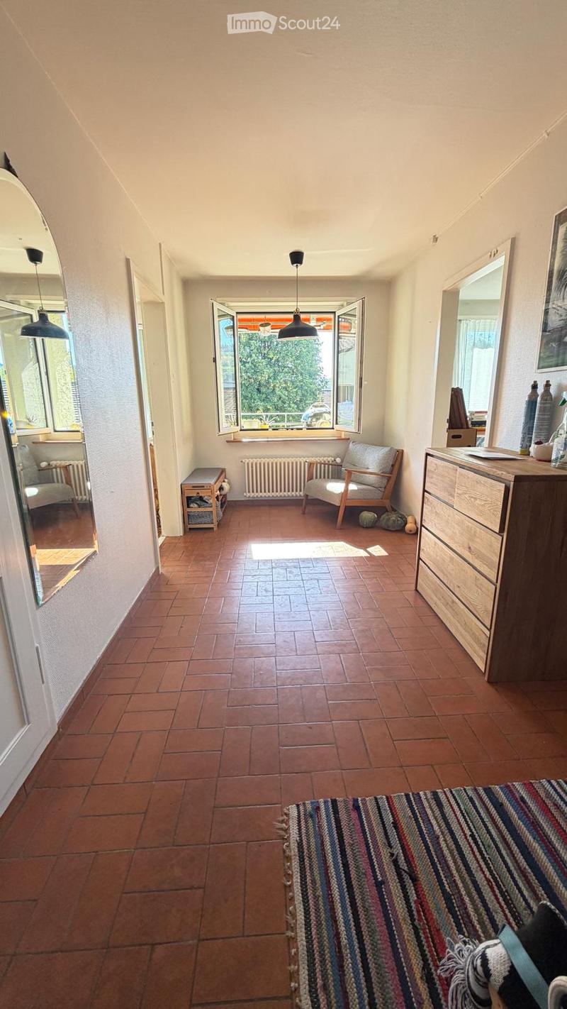 A cozy hallway with terracotta tiles, a colorful striped rug, a wooden chair, a small table, and a large mirror on the wall.