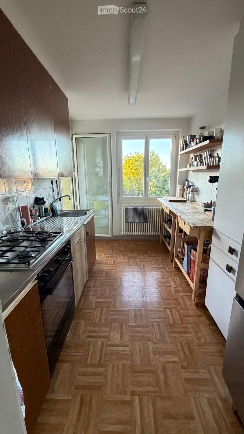 Modern kitchen with wooden herringbone flooring, fitted cabinets, a central island table, and a stove. Large windows provide natural light, and a radiator is visible beneath the windows.