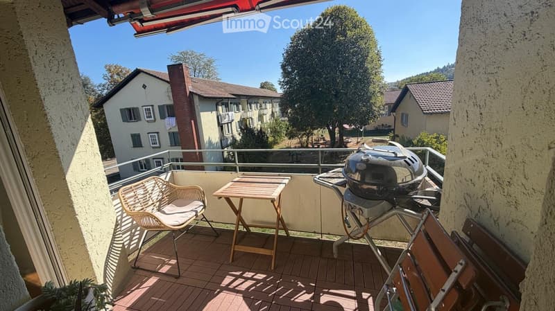 Balcony with wooden floor, wicker chair, small table, and grill, overlooking residential area with houses and trees.