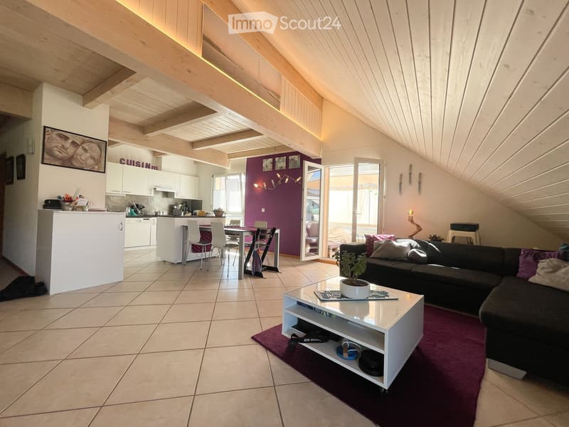 Open-plan living and dining area with exposed wooden beams, a black sectional sofa, a white coffee table, a potted plant, and a modern kitchen with white cabinets and a dishwasher.